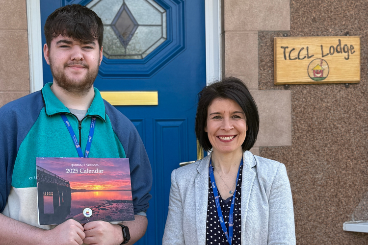 Two people outside a house with a sign saying TCCL Lodge, one is holding a calendar which was sold to raise money for the TCCL charity
